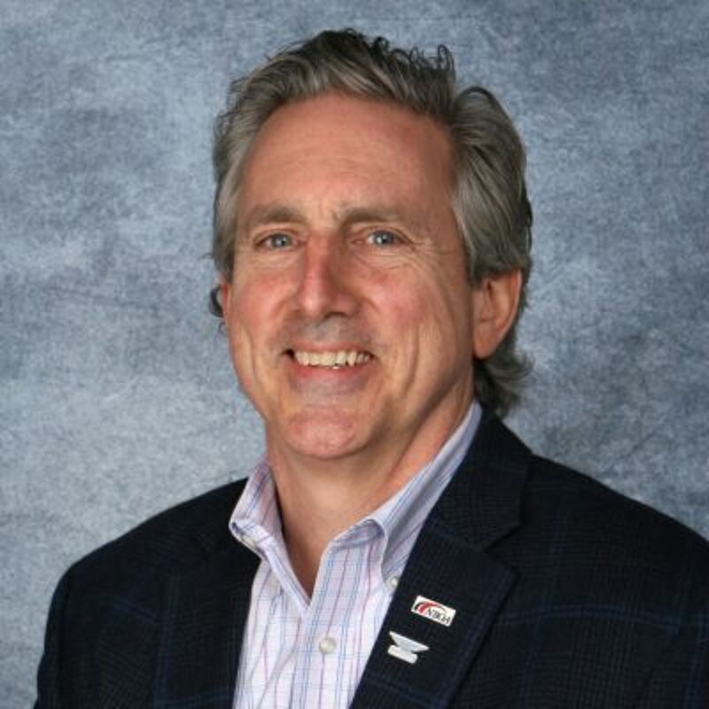 The image shows a middle-aged man with graying hair, smiling at the camera. He is wearing a blue blazer over a light blue and white striped shirt. The background is a blurry gray. He appears to be in a professional setting.
